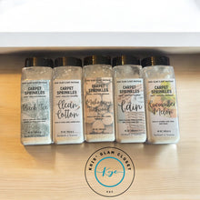 Load image into Gallery viewer, Five bottles of carpet sprinkle products arranged in a row on a light wooden surface, each labeled with different scents: Black Tea, Clean Cotton, Mahogany Teakwood, Gain, and Cucumber Melon. The bottles feature a chic design with a black lid and decorative fonts. A circular logo labeled "Kris's Glam Closet" is visible below the bottles.