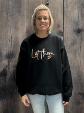 Load image into Gallery viewer, Woman with shoulder-length blonde hair wearing glasses, smiling and standing in front of a wooden background, dressed in a black sweatshirt featuring the text "Let them" in a stylish, light-colored font.