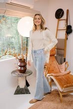 Load image into Gallery viewer, A woman stands in a bright, stylish room, smiling and wearing a light beige top, a white crocheted cardigan, and light blue flared jeans. She is near a large window with greenery outside and a standing lamp. A vase with branches sits on a round table beside her, and there is a comfortable chair with a tan cushion and a wooden ladder with hats in the background.