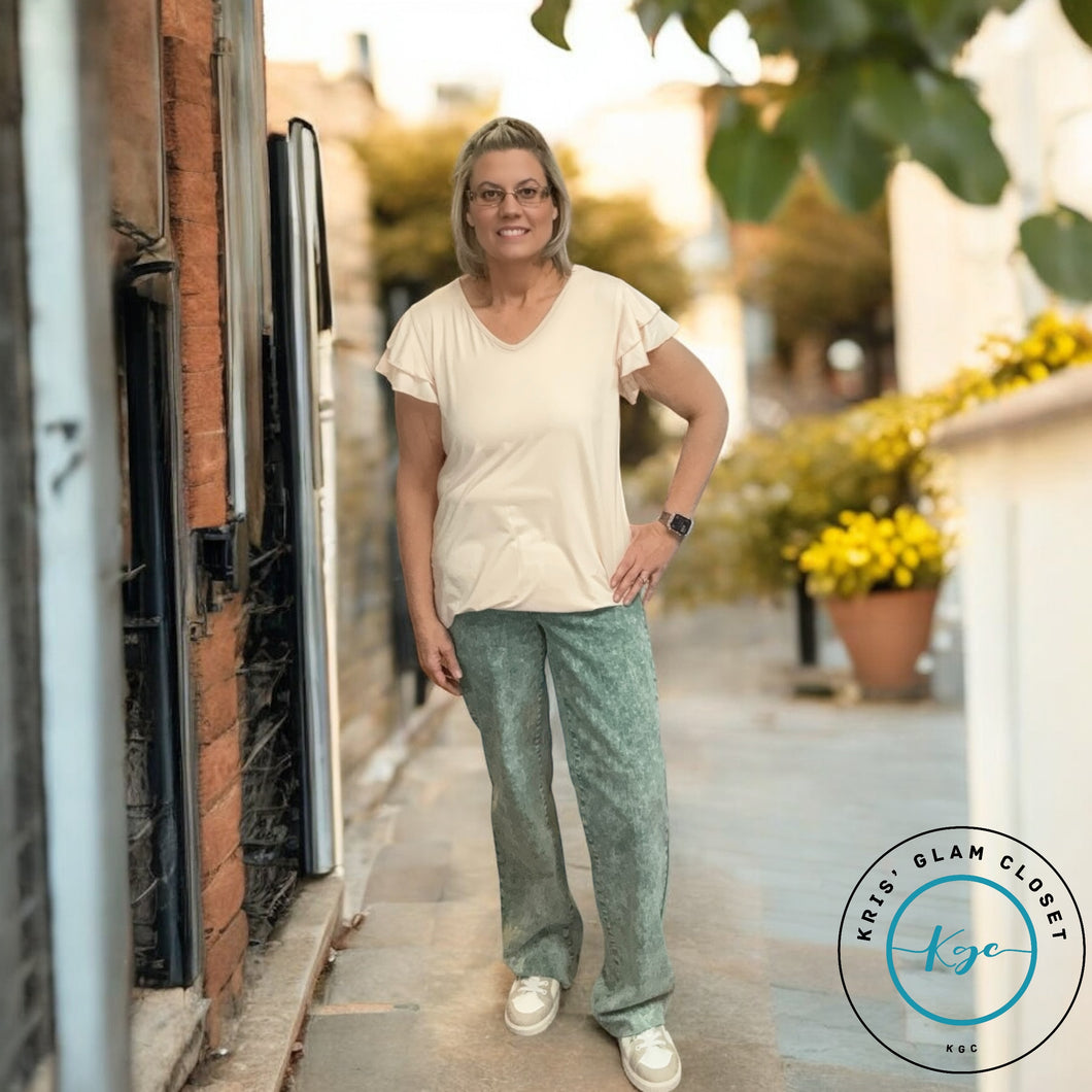 Woman standing on a pathway wearing a light beige v-neck t-shirt and green textured wide-leg pants, with white sneakers. Bright, sunny outdoor setting with potted yellow flowers in the background. Logo in the bottom right corner.